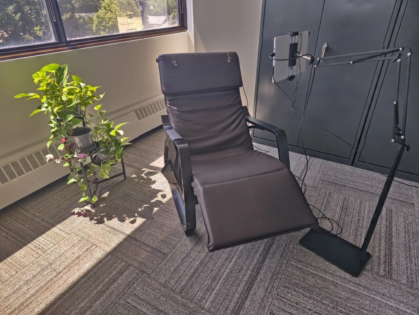 A brown leather reclining chair in our Burlington Office, with a tall potted plant with green leaves and pink flowers nearby, by a large window with sunlight streaming in, and a dark grey cabinet with a neurofeedback setup.