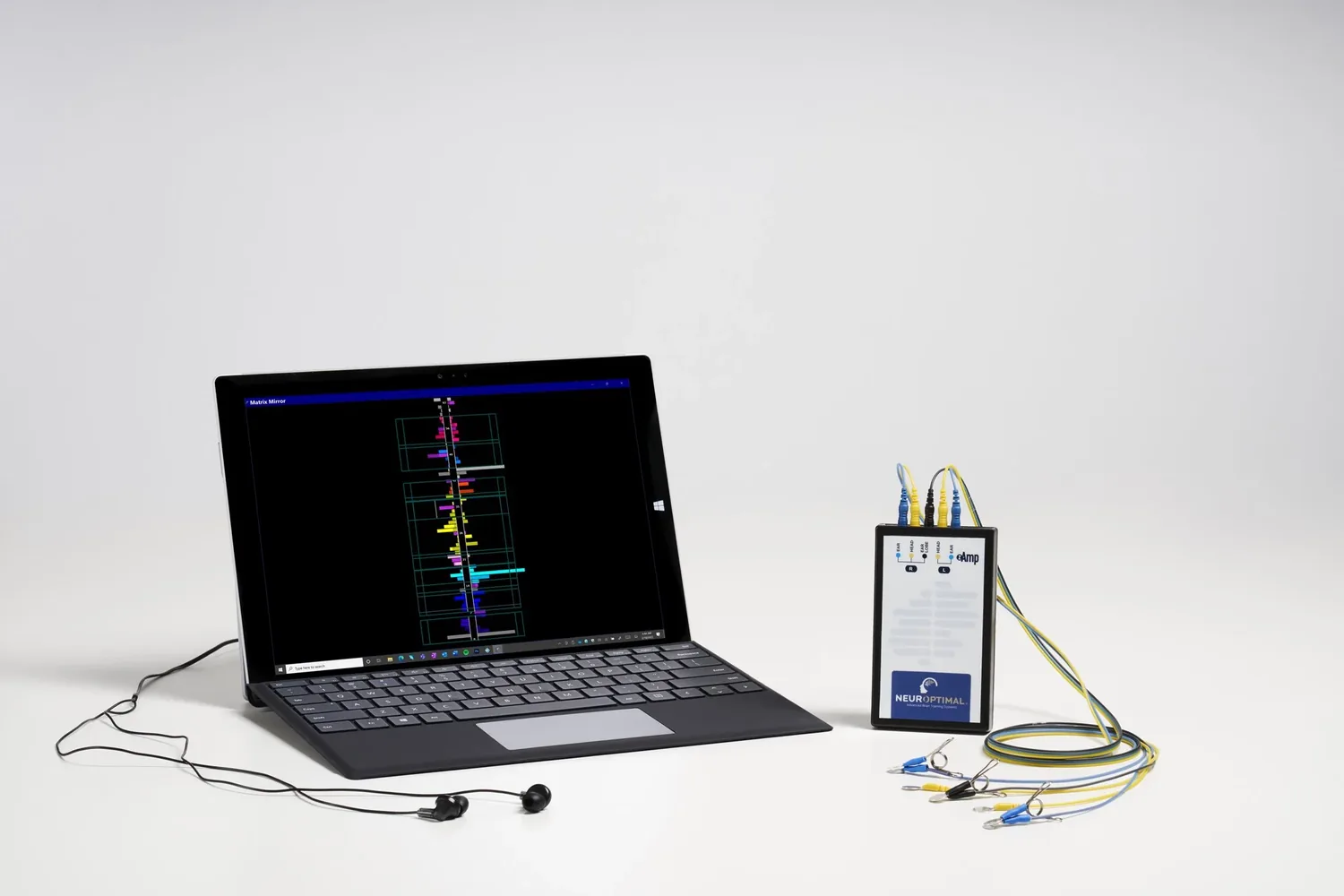 A NeurOptimal® Neurofeedback device displaying a colorful graph of EEG activity, connected to a small electronic device with neurofeedback sensors, and a pair of in-ear headphones placed on a white surface against a plain white background.